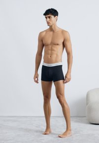 Czarne bokserki w wzory z białym pasem. Wykonane z miękkiego materiału, dopasowany krój, styl średniego stanu. Model stoi na jasnoszarym dywaniku.