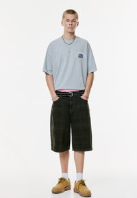 Ung mand står med hænderne i lommerne, iført en lysegrå oversized T-shirt med tekst, mørke knælange shorts og brune støvler med hvide sokker.