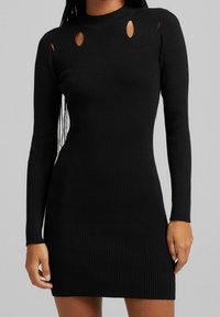 Svart ribbad stickad klänning med långa ärmar, med urklipp på axlarna och hög halsringning. Bodycon-passform framhäver silhuetten.