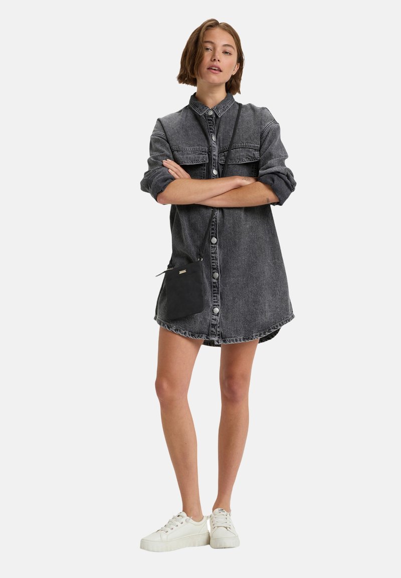 Vestido camisas de mezclilla en gris oscuro con cierre de botones, dos bolsillos en el pecho, mangas enrolladas y un pequeño bolso cruzado negro. Llevado con zapatillas blancas.