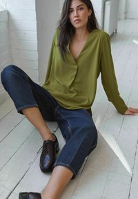 Un haut vert olive à manches longues avec un décolleté en V, associé à un jean en denim bleu court et des chaussures à lacets rouge foncé, sur un sol en bois blanc.