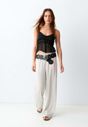 Jeune femme portant un caraco noir transparent, un pantalon large blanc, une grande ceinture noire circulaire et des sandales, debout devant un fond blanc.