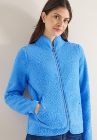 Femme aux longs cheveux bruns portant une veste en polaire bleu vif avec fermeture éclair et poches à pression, se tenant devant un mur clair uni.