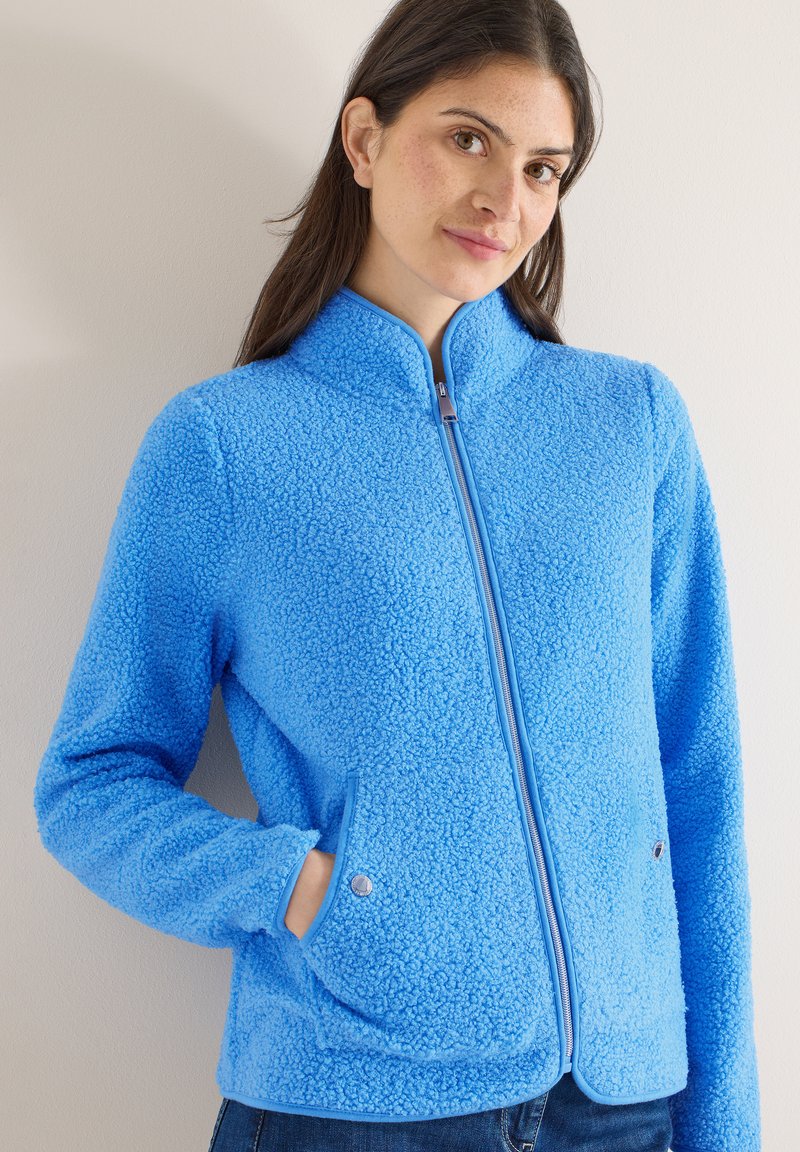 Femme aux longs cheveux bruns portant une veste en polaire bleu vif avec fermeture éclair et poches à pression, se tenant devant un mur clair uni.