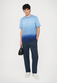 T-shirt à dégradé bleu avec des rayures horizontales, associée à un pantalon bleu marine et des baskets blanches. Le mannequin tient un petit sac à main noir.