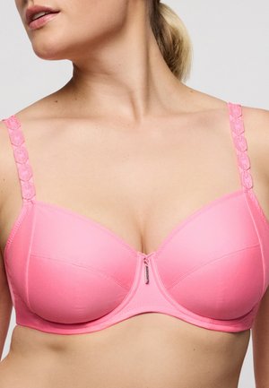 Vrouw draagt een roze gevoerde beha met decoratieve bandjes en een kleine metalen hanger in het midden aan de voorkant, haar nek naar de zijkant gedraaid.