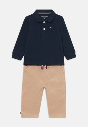 Marineblaues Langarmpolo mit einer Knopfleiste mit zwei Knöpfen und kleinem Logo, kombiniert mit beigefarbenen Sweatpants mit Kordelzug, die seitliche Taschen und kontrastierende Akzente aufweisen.