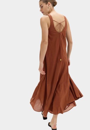 Vrouw draagt een mouwloze maxi-jurk in roestkleur met een open rug en strikdetail, lopend op zwarte schoenen met open hiel op een grijze achtergrond.