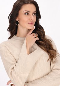 Pulover bej dintr-un material texturat, cu manșe late. Cercei închiși la culoare, inel cu un finisaj lucios. Model într-un cadru neutru.