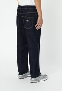 Dark blue denim pants with a loose fit, contrasting yellow stitching, and two back pockets. Paired with gray sneakers.