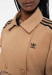 Jasnobrązowy płaszcz trench Adidas z czarnymi paskami na ramionach, dwurzędowy fason, duży kołnierz i logo po lewej stronie.