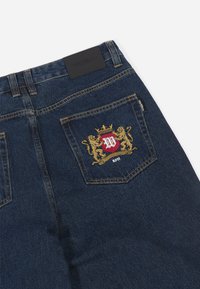Tamno plavi traperice s back džepom na kojem je zlatni i crveni izvezeni grb, kožni označivač na struku i žute šavove.