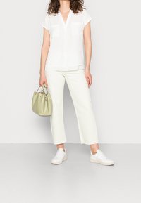Blouse bianca a maniche corte con scollatura a V e due tasche sul petto, abbinata a pantaloni cropped chiari e sneakers bianche. Tiene una borsa verde.