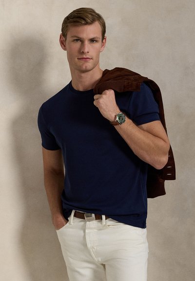 Moški v mornarsko modri majici in belih hlačah drži rjavo jakno čez ramo, nosi uro z zelenim številčnico in usnjenim paščkom.