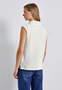 Weißes, ärmelloses Oberteil aus strukturiertem Strick mit einem hohen Kragen. Hat seitliche Schlitze am Saum und wird mit dunkelblauen Jeans kombiniert.