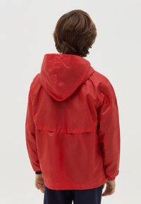 Giacca impermeabile rossa con cappuccio, polsini elasticizzati e flap posteriore. Texture liscia, vestibilità rilassata e design di lunghezza media senza hardware visibile.