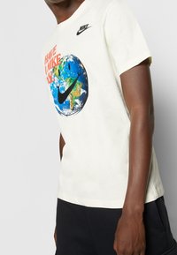 Nike puuvillane T-särk helesarnases värvitoonis, millel on graafika Maast ja naerunäo pildist. Sellel on must logo rinnal ja ümmargune kaelus.