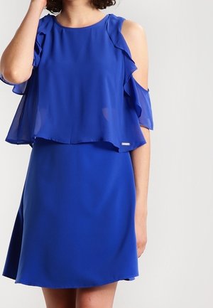 Femme portant une robe bleu royal à épaules dénudées avec des manches volantées et un haut fluide sur une jupe ajustée, debout devant un fond uni.