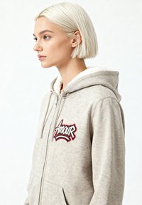 Hoodie zippé beige avec un intérieur texturé, présentant un logo "Amour" brodé en accents rouges et bleus sur la poitrine.