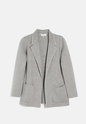 Kvinde i grå blazer og nederdel med hvid skjorte, der holder en brun håndtaske med kæderemme, iført guldsmykker og brune hæle.