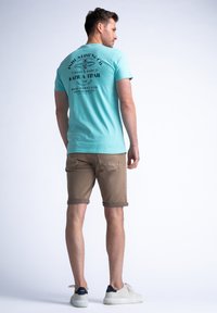 Camiseta de algodón color turquesa con un gráfico negro impreso en la parte trasera, combinada con pantalones cortos marrones enrollados y zapatillas blancas. Diseño minimalista.