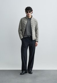 Geacă matlasată gri deschis, pulover în carouri alb-negru, pantaloni închiși la culoare, pantofi negri și ochelari de soare. Design geometric simplu.