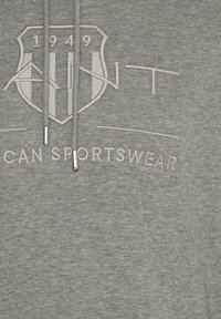 Felpa grigia con cappuccio caratterizzata da un logo a scudo con "1949" e "AMERICAN SPORTSWEAR" ricamati, cordini con estremità metalliche e un tessuto strutturato.