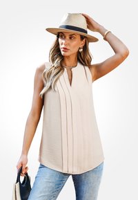 Beige mouwloos topje met verticale plooien, V-hals, zachte textuur, gecombineerd met lichtblauwe jeans en een strooien hoed. Kleine handtas in de hand.