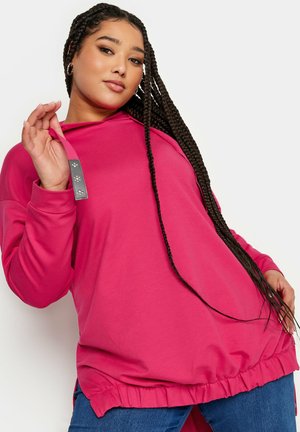 Sweat à capuche rose à manches longues, avec un ourlet festonné, des fentes sur les côtés et une capuche ajustée. Il présente un accent en matériel texturé au niveau du col.