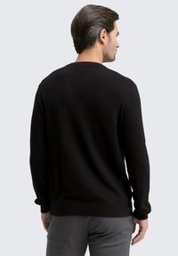 Mann, der sich mit dem Rücken zur Kamera in einem schlichten schwarzen langärmeligen Pullover und grauen Hosen vor einem hellgrauen Hintergrund steht.
