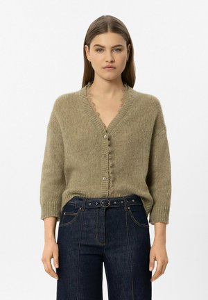 Frau trägt eine locker sitzende olivgrüne Strickjacke mit Knöpfen und dunkle Jeans mit hohem Bund und Gürtel, stehend vor einem einfarbigen Hintergrund.