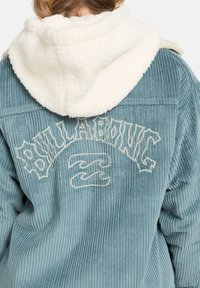 Blå fløjlsjakke med en cremet sherpa-fôret hætte. Har "BILLABONG" broderet i hvidt på ryggen med et bølgeikon nedenfor.