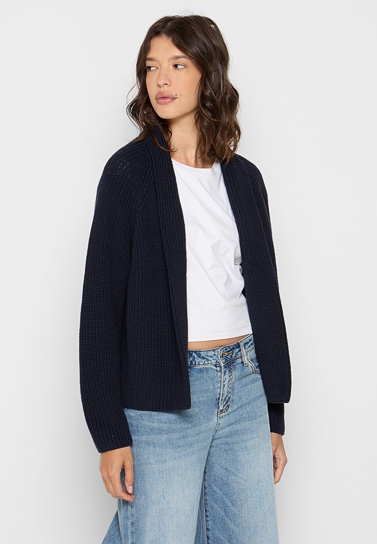 Marineblauer Strickcardigan, offenes Design, lange Ärmel, strukturierter Stoff. Getragen über einem weißen Crop-Top und hellblauen Jeans.