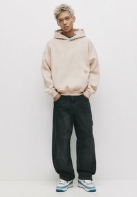 Felpa oversize beige con tasca frontale, abbinata a jeans scuri larghi e sneaker bianche con dettagli blu. Tessuto dalla consistenza morbida.