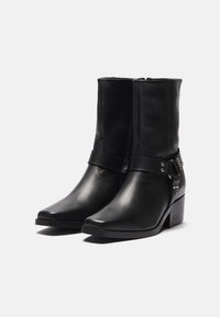 Zwarte leren enkelboots met een samengeperste neus, een lage blokhak, een zijrits en een decoratieve riem met metalen accenten.