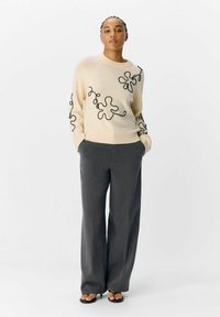 Maglione crema con ricami astratti neri sulle maniche, abbinato a pantaloni grigi a gamba larga e sandali neri. Texture liscia su tutto il capo.