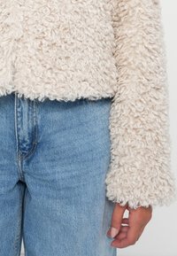 Giacca fuzzy color crema con un tessuto texturizzato e riccio e un design corto. Abbinata a jeans blu chiaro, che mostrano un look casual.