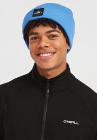 Beanie blu a coste con logo a patch nero. Indossato con una giacca nera con zip e logo bianco. Texture liscia e design aderente.