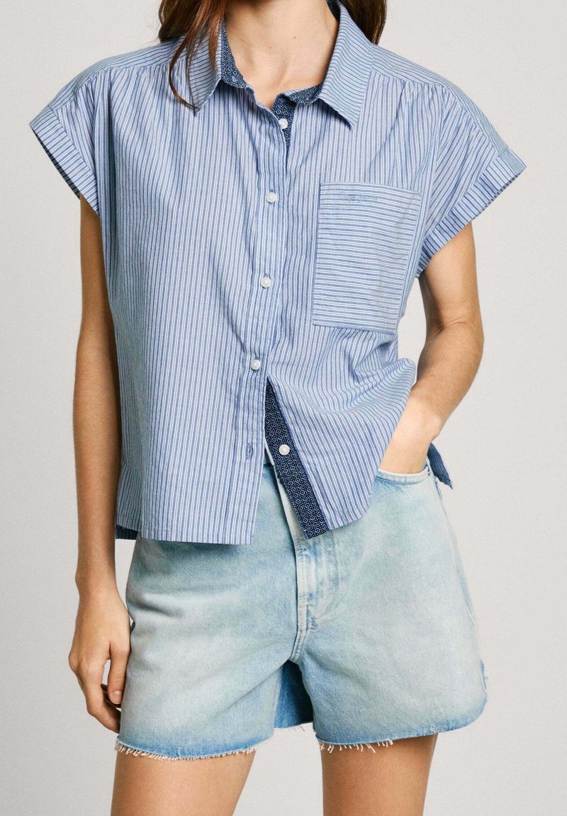 Femme portant une chemise à manches courtes rayée bleue avec poche poitrine et un short en denim clair aux bords effilochés, main dans la poche.