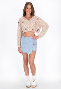 Junge Frau lächelt, trägt eine mehrfarbige, cropped Strickjacke, einen hellblauen Denim-Minirock, weiße Socken und weiße Sneakers, steht mit der Hand auf der Hüfte.