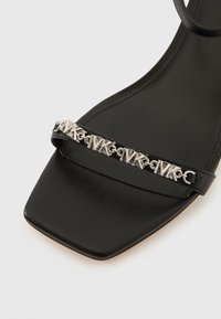 Μαύρο πέδιλο με τακούνι και τετράγωνη μύτη, με μεταλλικό λουράκι που φέρει λεπτομέρειες με λογότυπο και λεπτό λουράκι στον αστράγαλο. Ομαλή υφή δέρματος.