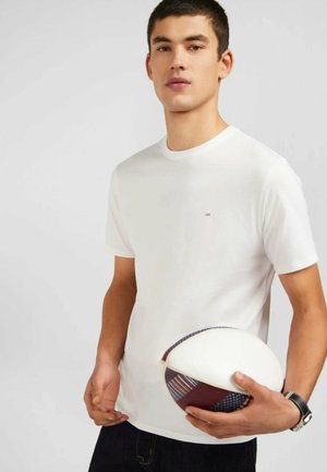 T-shirt en coton blanc avec un col rond et un petit logo violet, porté par une personne tenant un ballon de rugby à motifs multicolores.