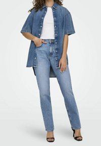 Camisa de mezclilla de manga corta en azul claro, con botón frontal y bolsillo en el pecho, combinada con jeans rectos en azul claro y tacones negros.