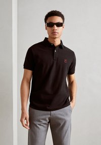 Schwarzes Baumwollpoloshirt mit Kragen und einem kleinen roten Logo auf der Brust, kombiniert mit grauen Hosen; das Model trägt dunkle Sonnenbrillen.