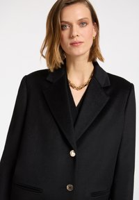 Blazer nero su misura con colletto a punte, tre bottoni dorati e un tessuto morbido e textured. Include tasche con patta e una scollatura a V sotto.