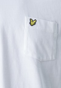 Hvit bomull T-skjorte med en liten, gul brodert ørn på en svart lomme. Glatt tekstur med et enkelt design.