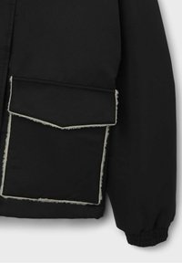 Svart jacka med en stor framficka med vit pälskant, elastiska ärmslut och en slät, matt tygkvalitet.