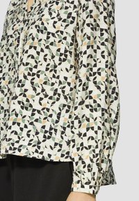 Blouse à motifs présentant des feuilles vertes, noires et beiges sur un fond clair. Manches longues avec des poignets élastiques et une coupe décontractée.