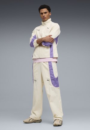 Ung mand står med armene krydset iført creme- og lilla Puma-træningsdragt og beige sneakers foran en ensfarvet grå baggrund.