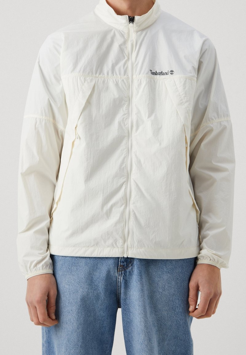 Homme portant une veste zippée Timberland blanche à manches longues et un jean bleu sur un fond uni.